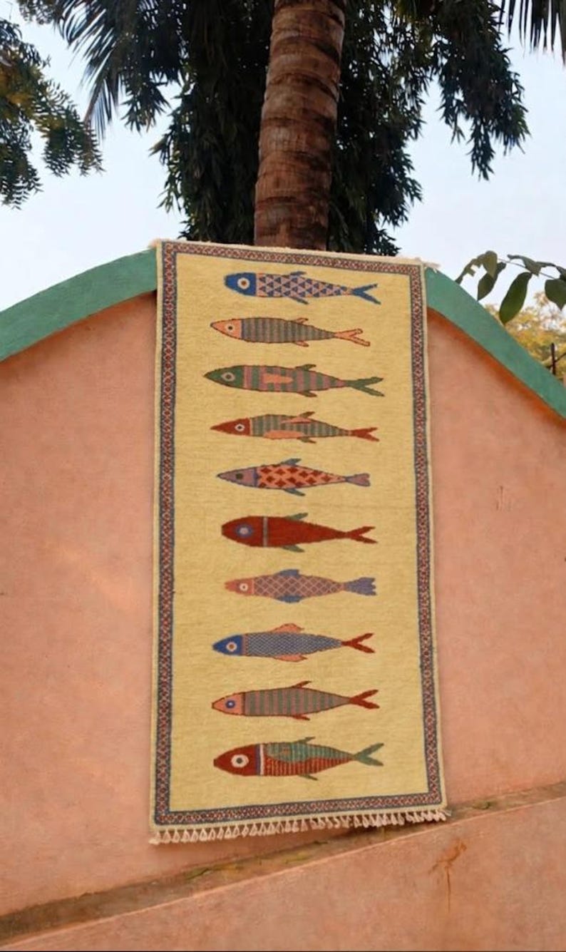 May include: A rectangular woven wall hanging featuring a light yellow background with a vertical row of ten stylized fish. Each fish displays a unique pattern and color combination, including shades of blue, red, green, and orange. The piece has a decorative border.