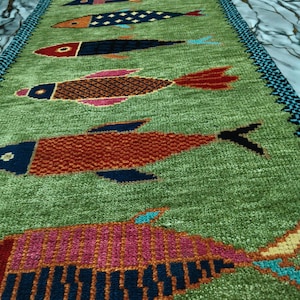 May include: A long, rectangular green runner rug featuring a repeating pattern of stylized fish in various colors including red, pink, orange, and dark blue. The rug has a textured pile and a dark blue and black patterned border along the edges.
