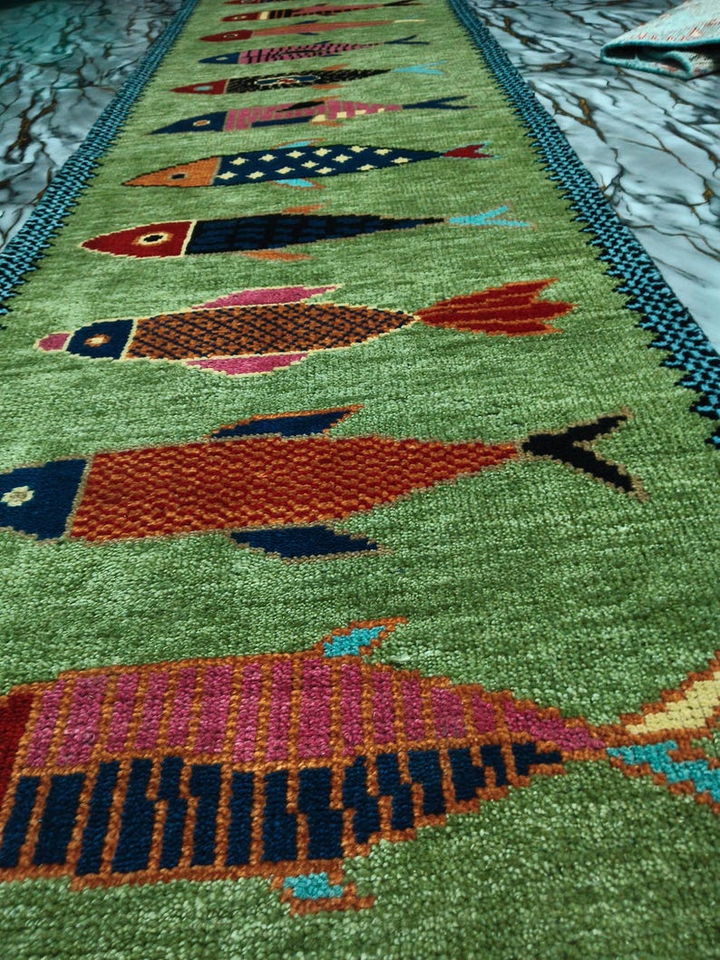 May include: A long, rectangular green runner rug featuring a repeating pattern of stylized fish in various colors including red, pink, orange, and dark blue. The rug has a textured pile and a dark blue and black patterned border along the edges.
