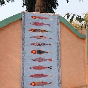 May include: A rectangular light blue woven wall hanging featuring a vertical pattern of ten stylized fish in various colors including red, pink, orange, and blue. The textile has a decorative dark border and white fringe at the top and bottom edges.