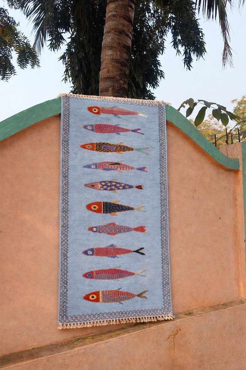 May include: A rectangular light blue woven wall hanging featuring a vertical pattern of ten stylized fish in various colors including red, pink, orange, and blue. The textile has a decorative dark border and white fringe at the top and bottom edges.