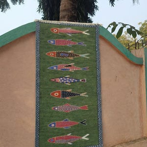May include: A rectangular green rug featuring a vertical row of nine stylized fish in various colors and patterns. The rug has a dark blue patterned border and white fringe at the top and bottom edges. The fish designs include red, blue, and yellow.