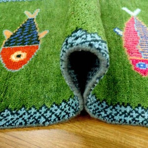 May include: A green area rug featuring a repeating pattern of stylized fish in orange, blue, and pink. The rug has a thick, tufted texture with a dark blue geometric border along the edges. One section of the rug is folded over to reveal the backing.