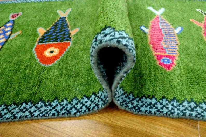 May include: A green area rug featuring a repeating pattern of stylized fish in orange, blue, and pink. The rug has a thick, tufted texture with a dark blue geometric border along the edges. One section of the rug is folded over to reveal the backing.