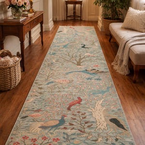 May include: A long, rectangular rug featuring a light blue backdrop with a detailed pattern of trees, birds, and floral elements. The rug has a cream-coloured fringe on both ends and is placed on a wooden floor.