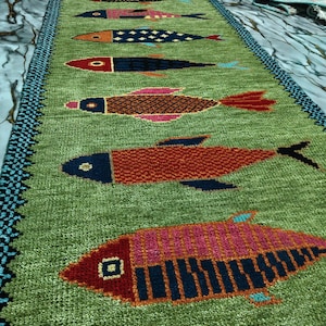 May include: A long, rectangular green table runner featuring a repeating pattern of stylized fish in various colors including red, blue, orange, and pink. The runner has a dark blue and black patterned border along the two long edges.