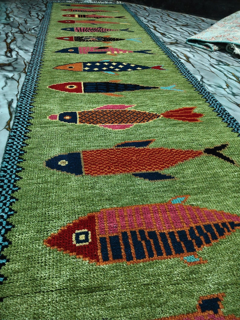 May include: A long, rectangular green table runner featuring a repeating pattern of stylized fish in various colors including red, blue, orange, and pink. The runner has a dark blue and black patterned border along the two long edges.