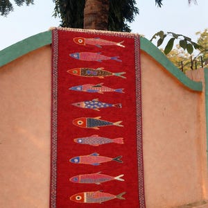May include: A rectangular red woven rug featuring a vertical column of ten stylized fish motifs. Each fish displays a unique pattern and color combination, including shades of blue, yellow, and pink. The rug has a decorative border and white fringe.