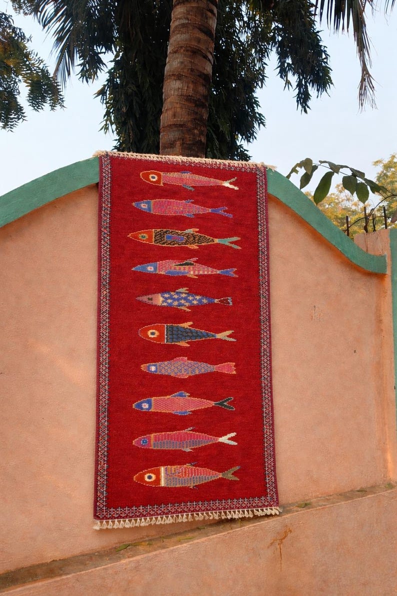 May include: A rectangular red woven rug featuring a vertical column of ten stylized fish motifs. Each fish displays a unique pattern and color combination, including shades of blue, yellow, and pink. The rug has a decorative border and white fringe.