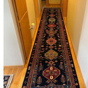 Op de afbeelding: Een lange, rechthoekige loper met een donkerblauwe achtergrond en ingewikkelde geometrische patronen in rood, groen en goud. Het tapijt heeft een gefranjerde rand en ligt in een gang met houten vloeren.
