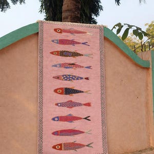 May include: A rectangular pink woven runner rug featuring a vertical pattern of ten stylized fish in various colors including red, blue, yellow, and purple. The rug has a dark geometric border and short white fringe at the top and bottom edges.