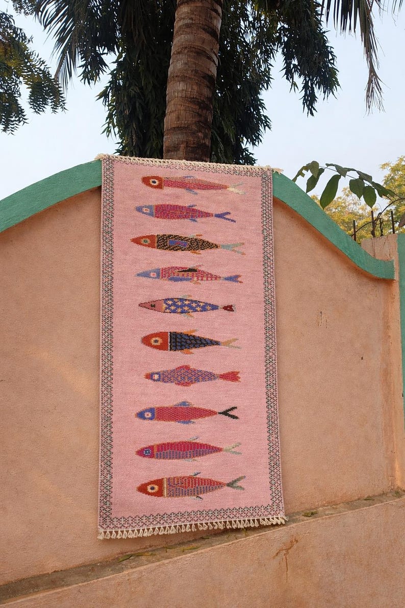 May include: A rectangular pink woven runner rug featuring a vertical pattern of ten stylized fish in various colors including red, blue, yellow, and purple. The rug has a dark geometric border and short white fringe at the top and bottom edges.