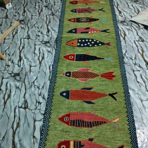 May include: A long, rectangular green runner rug featuring a vertical row of twelve stylized fish in various colors and patterns. The rug has a dark blue and black geometric border along both sides and a fringed edge at the top end.