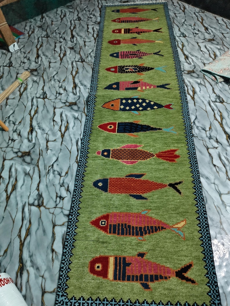 May include: A long, rectangular green runner rug featuring a vertical row of twelve stylized fish in various colors and patterns. The rug has a dark blue and black geometric border along both sides and a fringed edge at the top end.