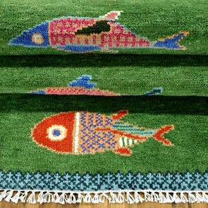 May include: A green rectangular rug featuring a repeating fish pattern in shades of red, blue, and yellow. The rug has a dark blue geometric border and white fringe along the bottom edge. The surface has a thick, textured pile construction.