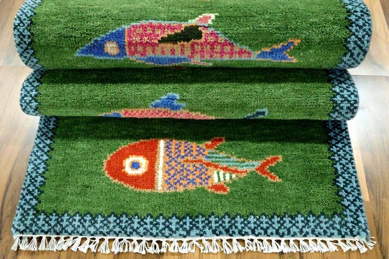 May include: A green rectangular rug featuring a repeating fish pattern in shades of red, blue, and yellow. The rug has a dark blue geometric border and white fringe along the bottom edge. The surface has a thick, textured pile construction.