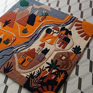May include: A rectangular tufted rug featuring a colorful landscape design with mountains, a winding river, small houses, trees, and sun motifs in shades of orange, blue, brown, and cream. The textured surface displays a detailed graphic scene.