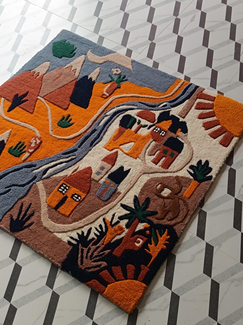 May include: A rectangular tufted rug featuring a colorful landscape design with mountains, a winding river, small houses, trees, and sun motifs in shades of orange, blue, brown, and cream. The textured surface displays a detailed graphic scene.