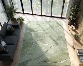 May include: A large, rectangular green rug with a leaf vein design. The rug features varying shades of green and white, creating a textured, natural look. The rug is placed on a wooden floor in a room with large windows and a view of trees.