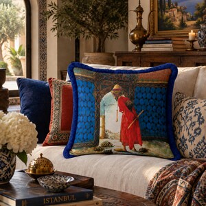 May include: A square decorative throw pillow featuring a vibrant illustration of a person in a red robe and yellow turban standing in a blue arched doorway, surrounded by small tortoises, finished with a thick, textured royal blue fringe border.