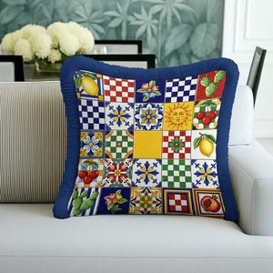 May include: Square decorative pillow with a vibrant patchwork design featuring lemons, cherries, flowers, and geometric patterns. The pillow has a blue fringe border and is placed on a white sofa.