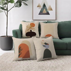 Könnte beinhalten: Vier quadratische Kissen mit abstrakten Designs in Orange, Beige und Schwarz liegen auf einem grünen Sofa. Die Kissen zeigen geometrische Formen und botanische Motive. Ein gerahmtes Kunstwerk hängt über dem Sofa.