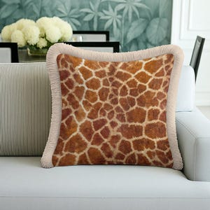Könnte beinhalten: Quadratisches Zierkissen mit Giraffenmuster in Braun- und Beigetönen. Es hat einen beigen Fransenbesatz und liegt auf einem weißen Sofa. Dieses Kissen verleiht einem Wohnzimmer einen Hauch von Safari-Dekor. Maße ca. 40 cm x 40 cm.