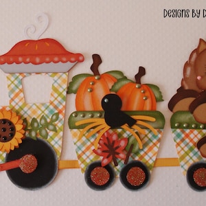 May include: A paper craft train decoration featuring a pie on the engine, pumpkins in a cargo car, and a brown squirrel in the caboose. The train is decorated with a sunflower, plaid patterns, and autumn leaves. Text reads DESIGNS BY DEBANDERSON651.