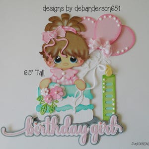 May include: A paper cutout of a birthday girl holding a cake, with pink balloons and a candle. The image is 6.5 inches tall. The words "birthday girl" are written in cursive. The design includes pink, blue, green, and brown colors.