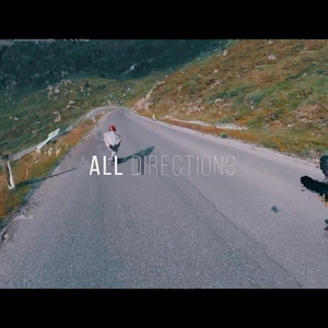 May include: A scenic shot of a winding mountain road with two skateboarders. One is riding down the road, and the other is performing a trick. The text "ALL DIRECTIONS" is overlaid on the road.