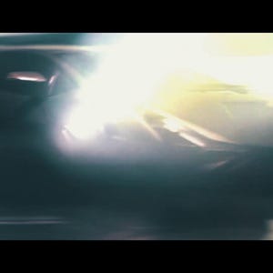 May include: A dark, abstract image featuring a car's headlight, emitting a bright, intense light. The car's body is partially visible, with a sleek, aerodynamic design. The overall aesthetic is cinematic and dramatic.