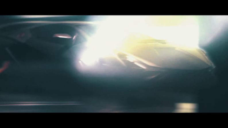 May include: A dark, abstract image featuring a car's headlight, emitting a bright, intense light. The car's body is partially visible, with a sleek, aerodynamic design. The overall aesthetic is cinematic and dramatic.