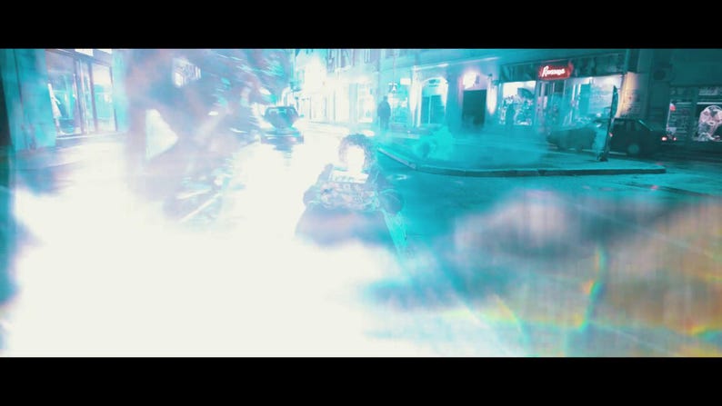 May include: A nighttime street scene with a bright, glowing light source in the foreground. The scene is dominated by a bright, white light with blue and teal hues. Buildings and figures are visible in the background.