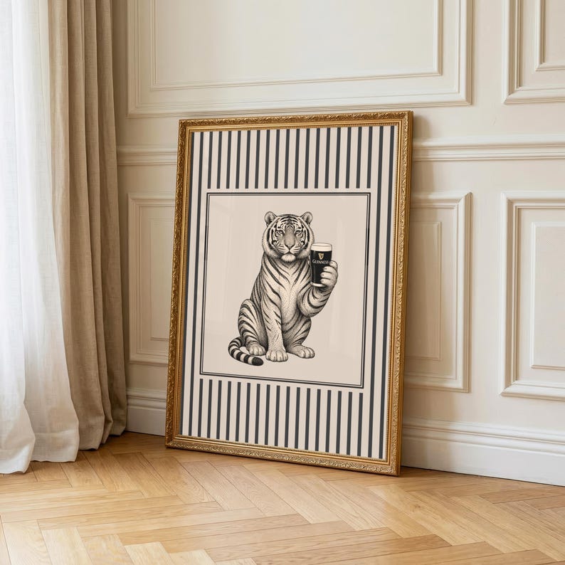 May include: A framed art print depicting a tiger holding a pint of Guinness. The black and white illustration has a gold frame and a striped border. The print is displayed on a light beige background. The artwork is a unique piece.