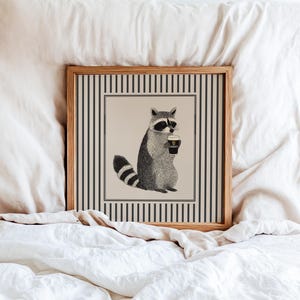 Op de afbeelding: Een ingelijste prent met een wasbeer die een pint Guinness vasthoudt. De wasbeer is afgebeeld in zwart-wit, met een gestreepte staart en masker. De prent heeft een gestreepte rand en een houten lijst, op een wit bed.