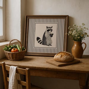 Op de afbeelding: Een ingelijste illustratie van een wasbeer met een koffiekopje, tegen een gestreepte achtergrond. De afbeelding staat op een houten tafel, samen met een brood, een groentemand en een vaas met bloemen.