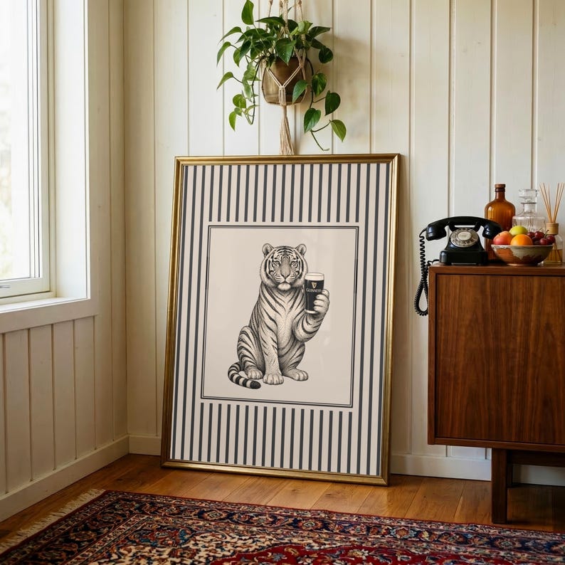 May include: A framed art print depicting a tiger holding a pint of Guinness. The monochrome illustration is set against a cream backdrop, framed with a gold border. The background features black and white vertical stripes. The artwork is displayed indoors.