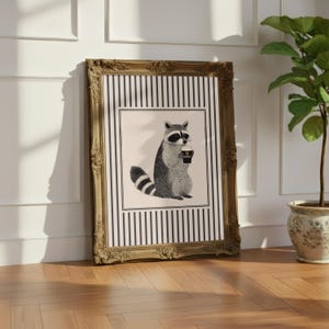 Op de afbeelding: Een ingelijste kunstdruk met een wasbeer die een kop koffie vasthoudt. Het kunstwerk staat tegen een gestreepte achtergrond en is ingelijst in een sierlijke gouden lijst. De print leunt tegen een witte muur en een houten vloer.