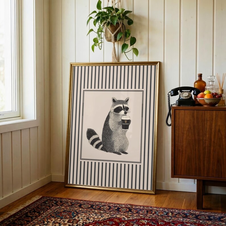 Op de afbeelding: Een ingelijste kunstprint met een wasbeer die een koffiekopje vasthoudt. De zwart-wit illustratie staat op een cr&egrave;mekleurige achtergrond, omlijst door een gestreepte rand en een goudkleurige lijst. De print leunt tegen een witte muur.