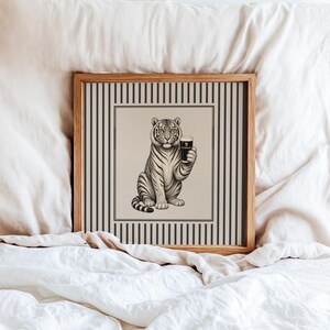 May include: A framed print depicting a tiger holding a pint of Guinness. The black and white illustration is set against a beige backdrop with a striped border. The frame is made of light-coloured wood, adding a classic touch.