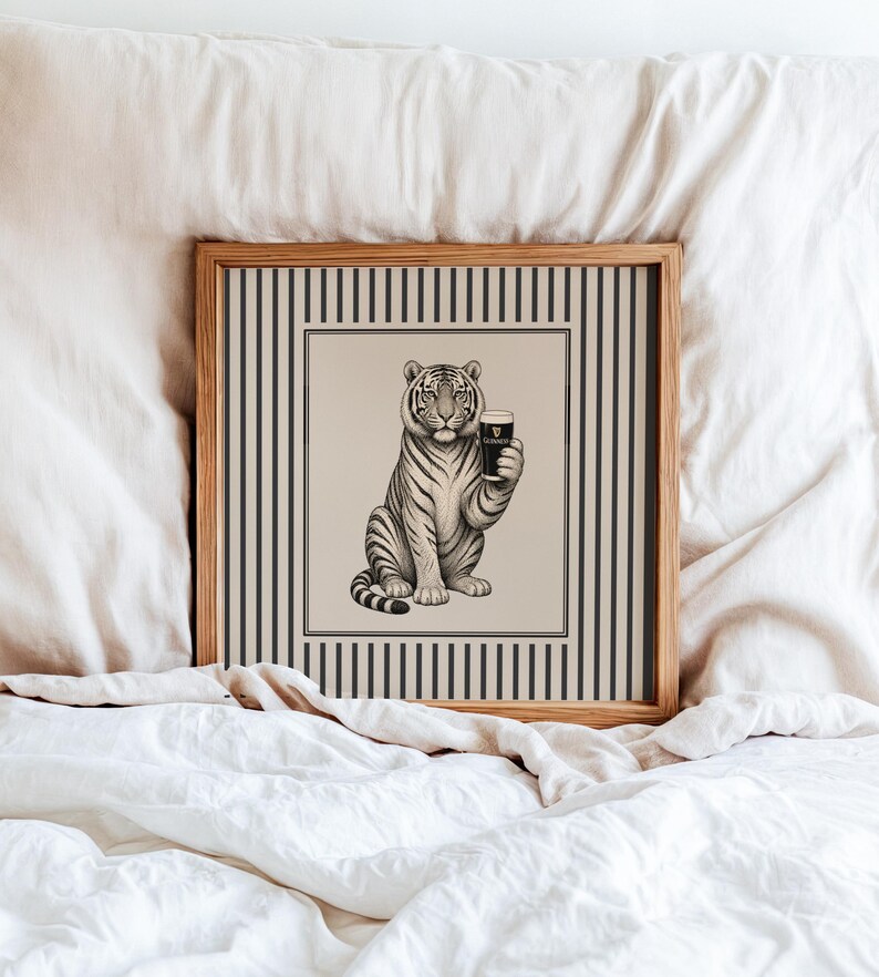 May include: A framed print depicting a tiger holding a pint of Guinness. The black and white illustration is set against a beige backdrop with a striped border. The frame is made of light-coloured wood, adding a classic touch.