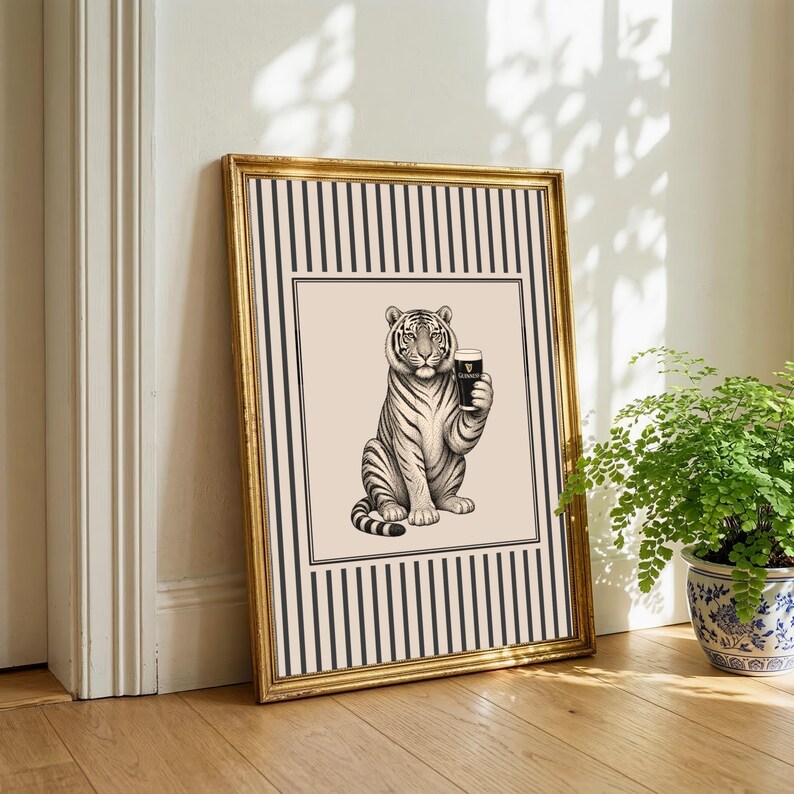 May include: A framed art print showing a tiger holding a pint of stout. The black and white tiger is set against a cream background with a striped border. The gold frame adds a touch of elegance to the artwork. The print is in a gold frame.