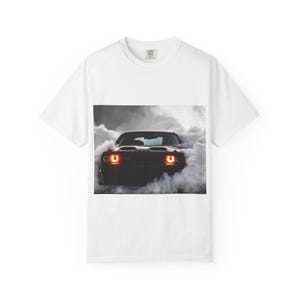 May include: White t-shirt featuring a graphic of a black muscle car engulfed in smoke. The car's headlights are illuminated, and the image is set against a backdrop of billowing gray smoke. The shirt has a crew neck and short sleeves.