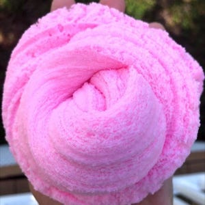 以下が含まれることがあります： 明るいピンク色のスライムの塊のクローズアップ。柔らかく、質感があります。スライムは渦巻き状に渦巻いており、隆起とふわふわした外観が見えます。背景はぼやけており、屋外環境を示唆しています。