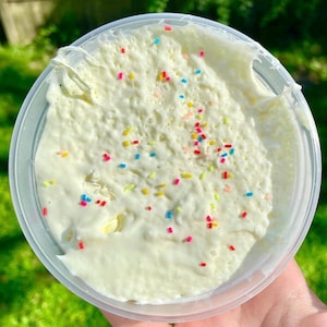 Op de afbeelding: Een bakje crème-witte slijm met kleurrijke sprinkles. De slijm heeft een textuur, romige uitstraling en zit in een doorzichtige plastic bak. De sprinkles zijn rood, geel, blauw en roze.