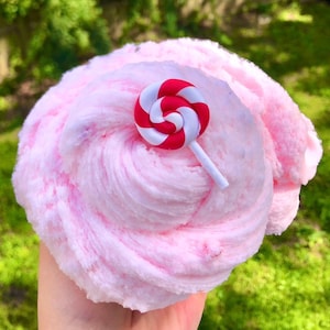 May include: A close-up of pink slime with a swirl design, decorated with a red and white lollipop. The slime has a soft, fluffy texture. The lollipop is a miniature, decorative element. The background is a blurred green, suggesting a garden.