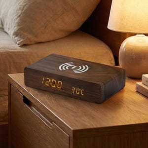 May include: A dark wood wireless charging alarm clock displaying the time and temperature. The clock has a rectangular shape with rounded edges and a white wireless charging symbol on top. The time reads 12:00 and the temperature is 30°C.