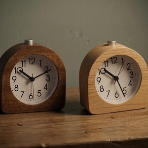 Puede incluir: Dos relojes despertadores de madera, uno oscuro y otro claro. Ambos tienen esferas blancas con números y manecillas negras. Los relojes miden unos 10 cm de alto y están sobre una superficie de madera.