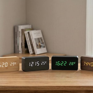 May include: Four rectangular digital clocks with wooden frames are displayed on a wooden table. The clocks show the time and temperature in degrees Celsius. The first clock is light-coloured wood, the second is dark grey, the third is black, and the fourth is brown.
