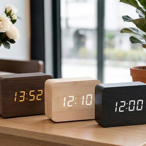 Può includere: Tre orologi digitali in legno marrone, legno chiaro e nero, che mostrano l'ora. L'orologio marrone mostra 13:52 in ambra, l'orologio in legno chiaro mostra 12:10 in bianco e l'orologio nero mostra 12:00 in bianco. Un vaso di fiori bianchi e una pianta in vaso sono sullo sfondo.